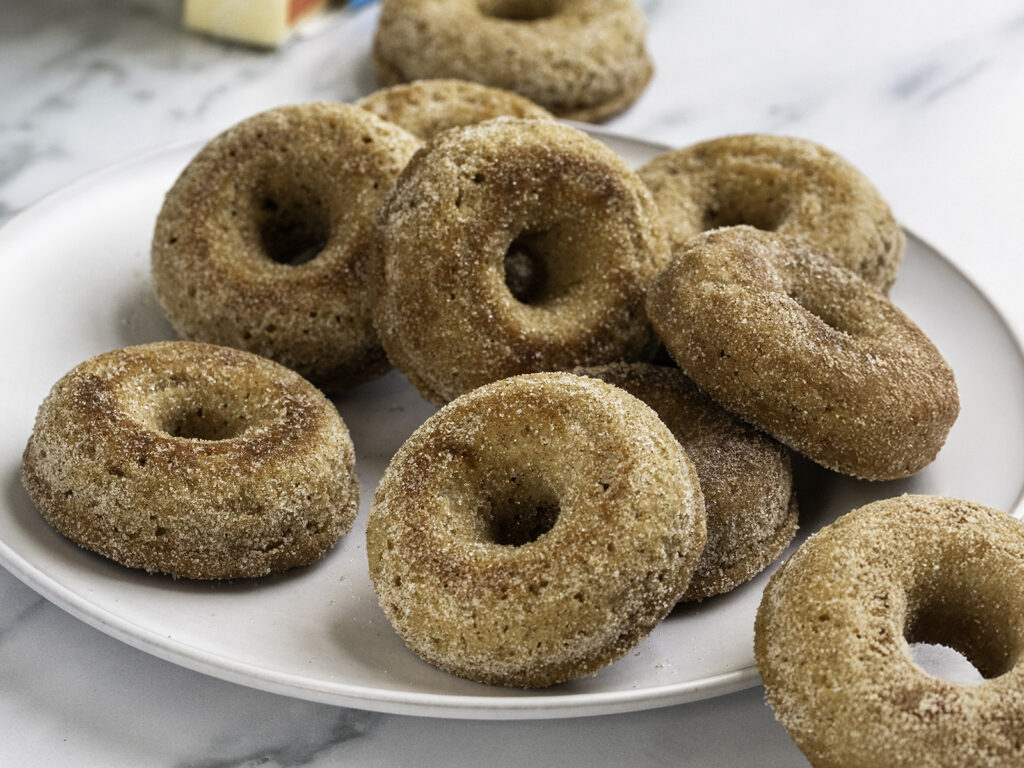 A plate of homemade buttermilk apple cider donuts coated in a cinnamon sugar mixture, stacked closely together on a white dish.