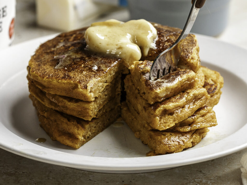 Stack of fluffy pumpkin cottage cheese pancakes on a white plate, topped with melting butter and syrup, with a fork cutting into the stack to reveal the soft, moist texture inside.