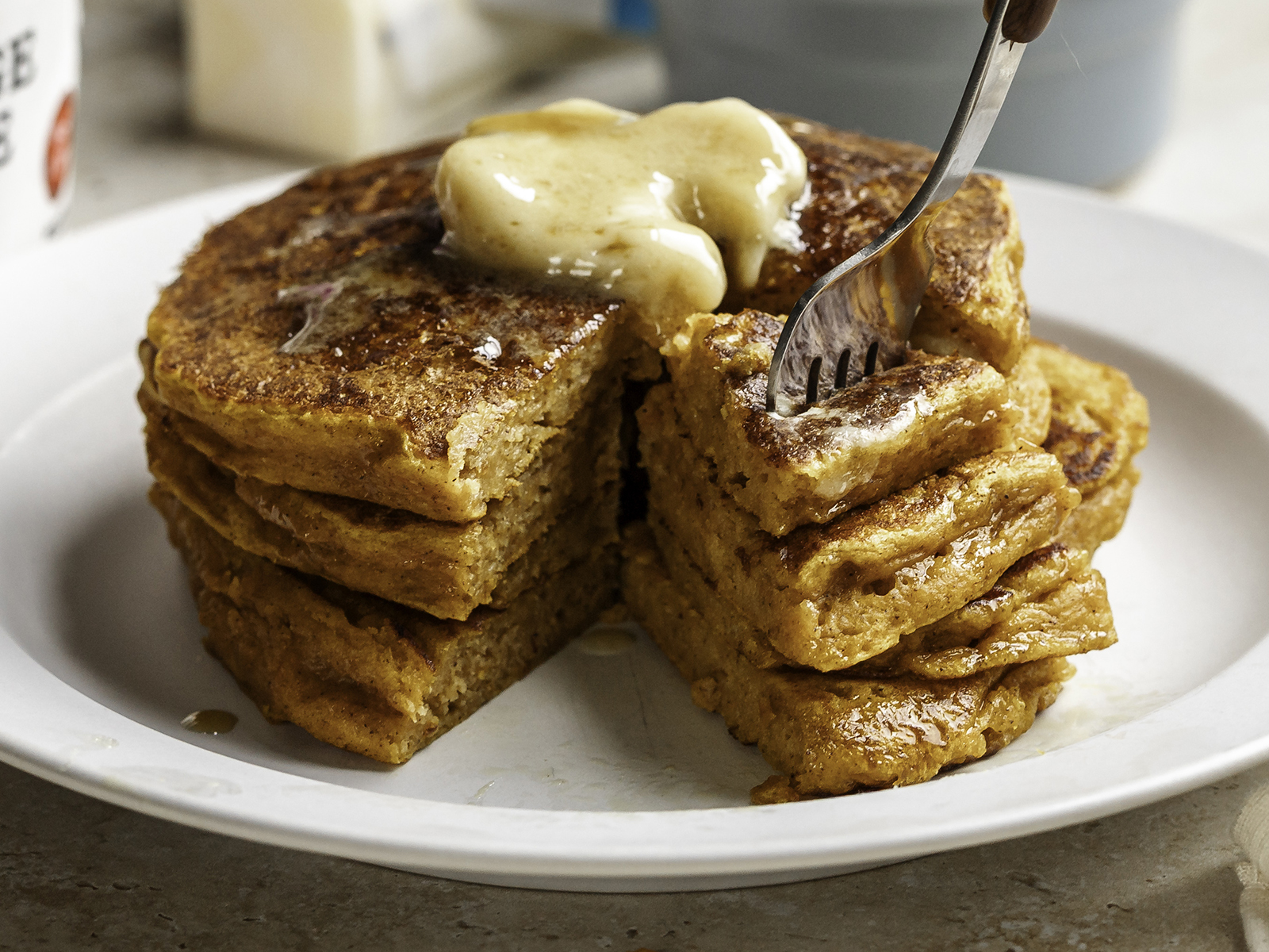 Stack of fluffy pumpkin cottage cheese pancakes on a white plate, topped with melting butter and syrup, with a fork cutting into the stack to reveal the soft, moist texture inside.