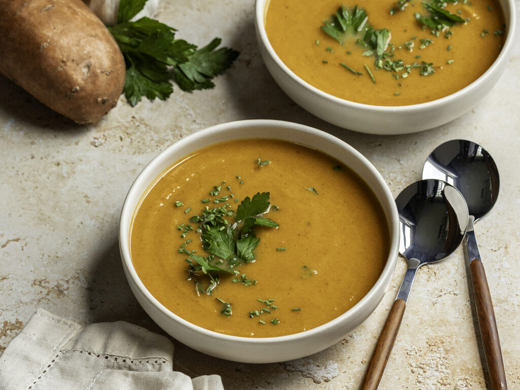 Two bowls of creamy roasted fall vegetable soup topped with fresh parsley, set on a light stone surface with spoons beside them and a sweet potato and parsley in the background.