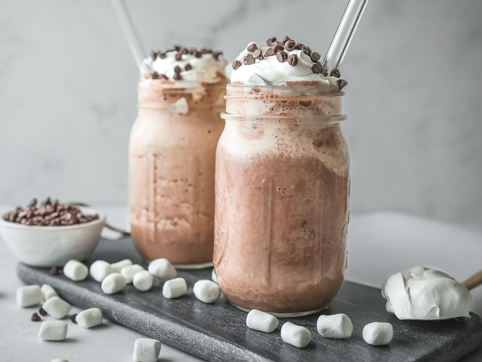 Two mason jars filled with frozen mint hot chocolate topped with whipped cream and mini chocolate chips, served with glass straws and surrounded by mini marshmallows on a dark slate board.