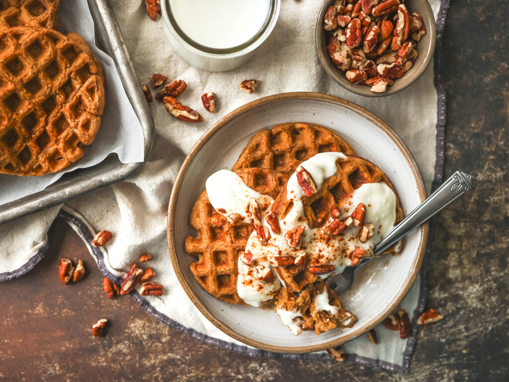 Plate of golden gingerbread waffles topped with creamy drizzle and chopped pecans, served with a glass of milk nearby.