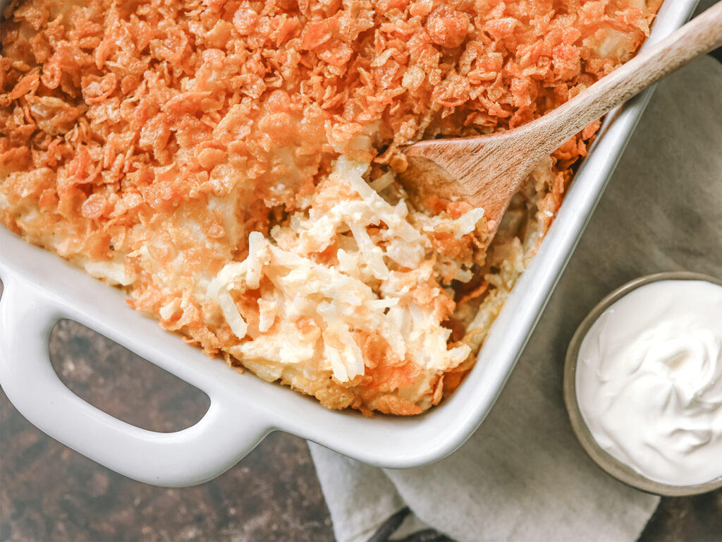 Creamy hash brown casserole topped with a crispy cornflake crust, served with a side of sour cream in a white baking dish.