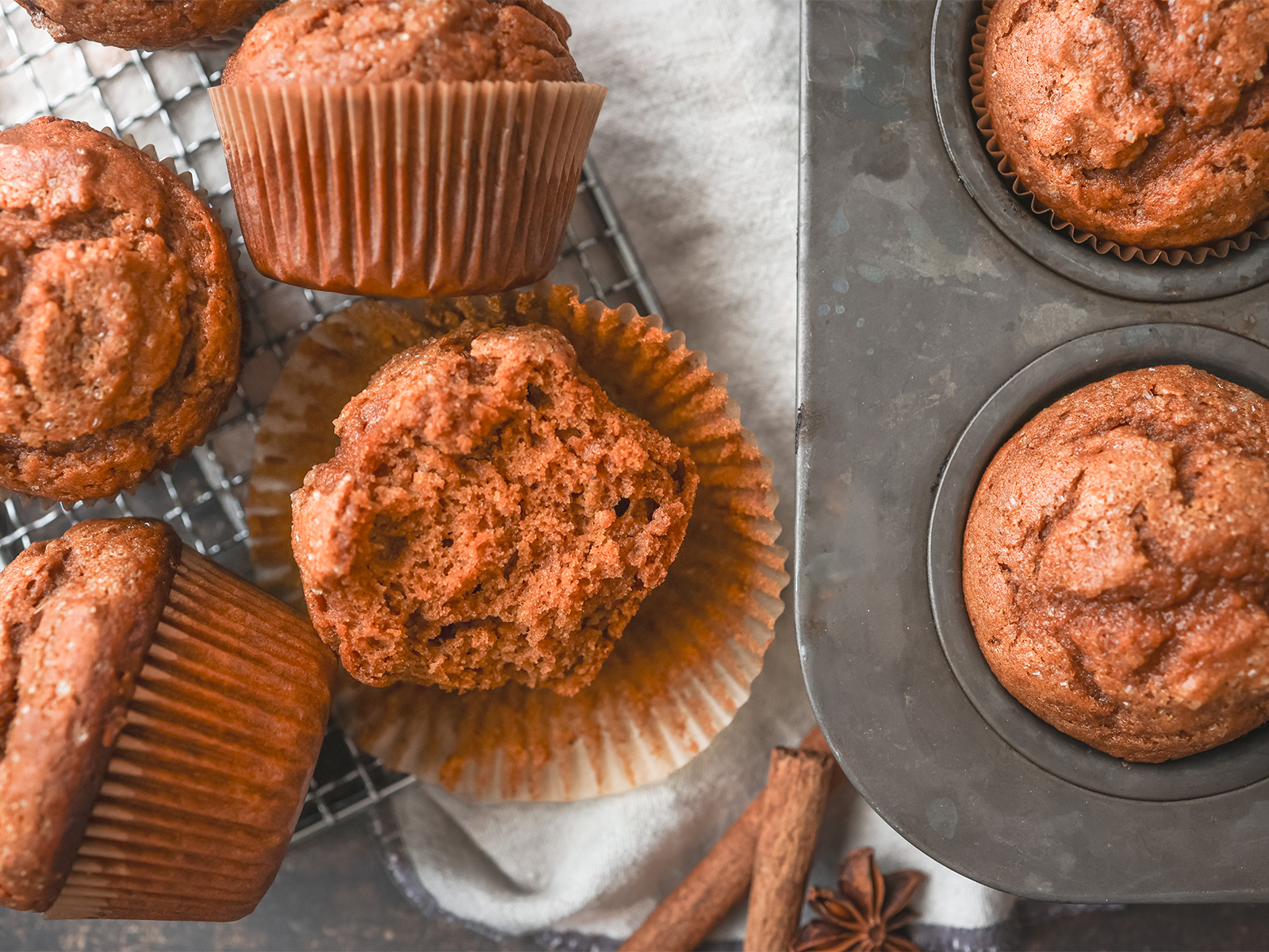 Freshly baked pumpkin spice muffins in a metal muffin tin and on a cooling rack, with one muffin cut open to show its soft, moist texture.