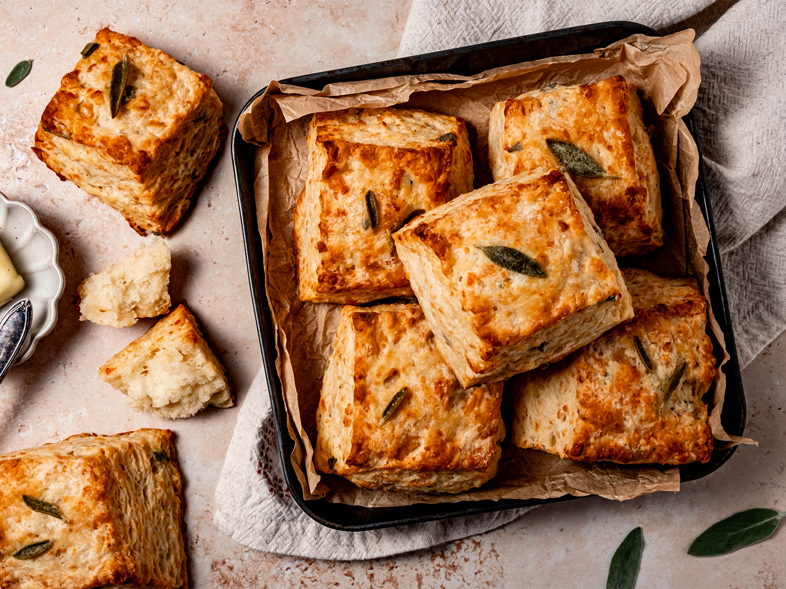 Golden, flaky buttermilk sage biscuits stacked in a parchment-lined pan with butter and sage leaves on the side.