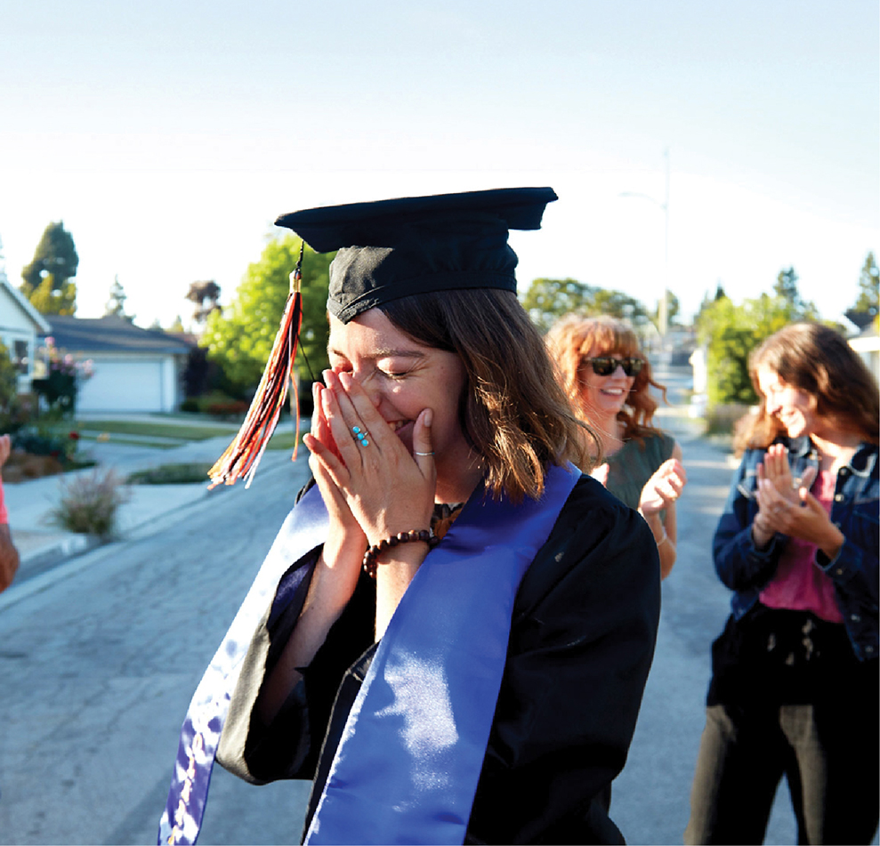 A young graduate wearing a cap and gown covers her face with her hands while crying with joy as people behind her applaud outdoors.