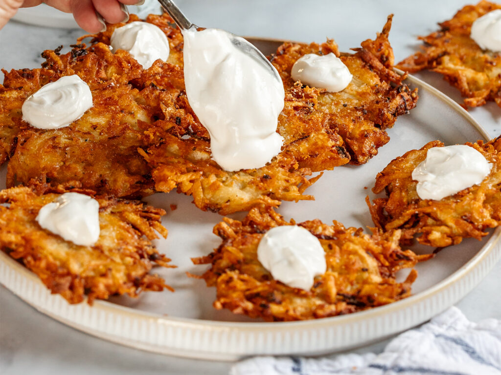 Close-up of crispy cheesy latkes on a plate, each topped with sour cream, as a spoon adds a generous dollop to one latke, highlighting the golden, crunchy texture.