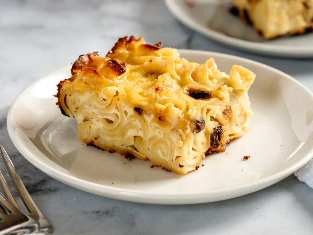 A slice of baked noodle kugel on a white plate, showing tender noodles with a lightly golden, caramelized top and creamy interior.