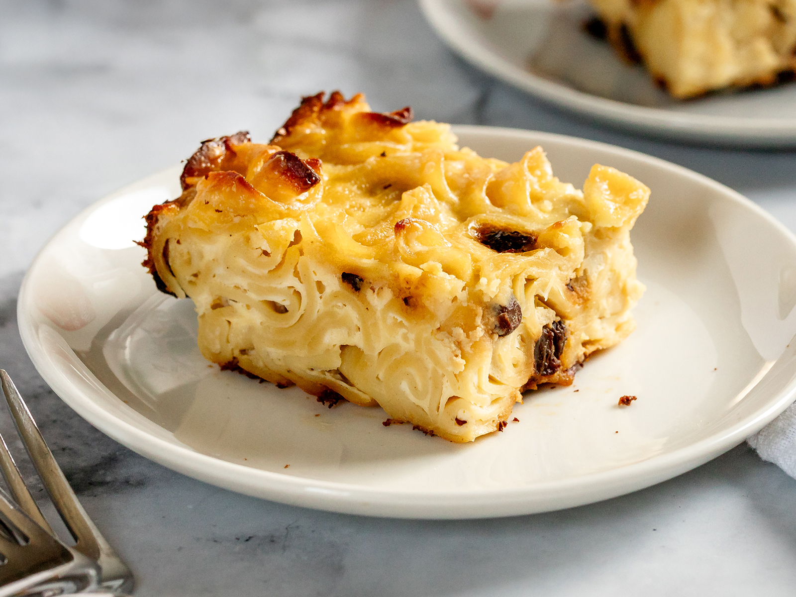 A slice of baked noodle kugel on a white plate, showing tender noodles with a lightly golden, caramelized top and creamy interior.