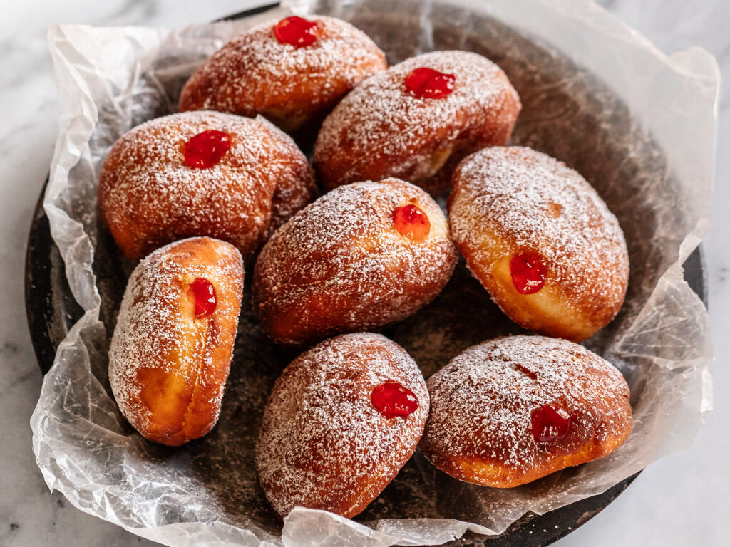A dark bowl lined with parchment paper containing eight jelly-filled fried doughnuts dusted with powdered sugar, each topped with a bright red jelly center.