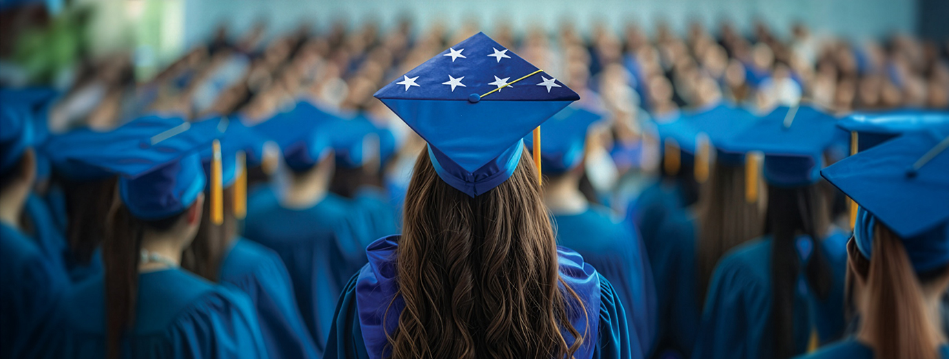 A graduate seen from behind wears a blue cap decorated with white stars while standing among rows of graduates in matching blue gowns at a commencement ceremony