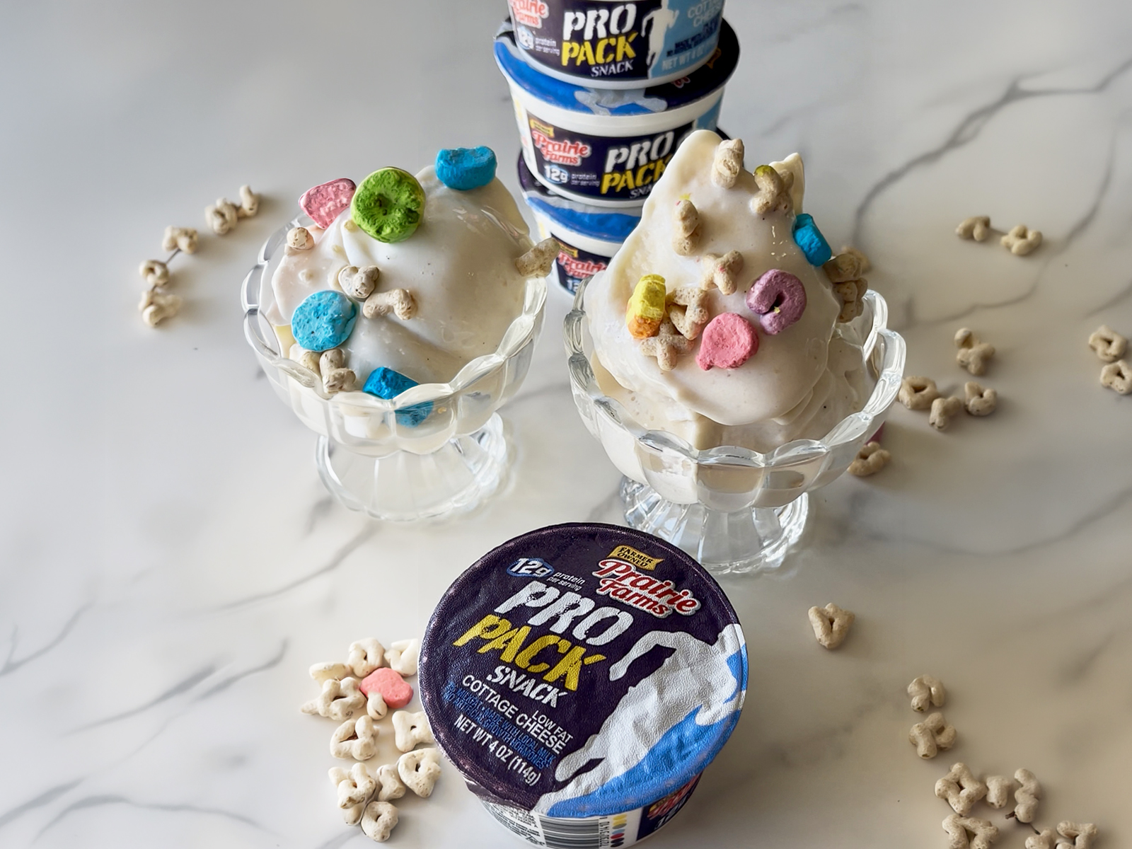 Two dessert bowls filled with creamy cottage cheese ice cream topped with marshmallow cereal pieces, with cottage cheese snack cups stacked in the background and cereal scattered on the surface.