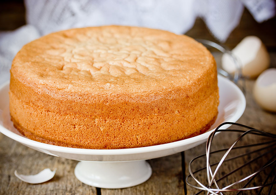Plain golden sponge cake on a white cake stand, with a whisk nearby.
