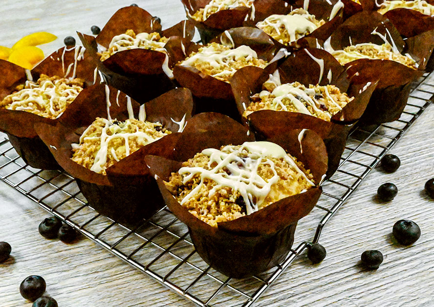 Muffins in brown paper liners on a cooling rack, topped with white icing and crumbs, with blueberries nearby.