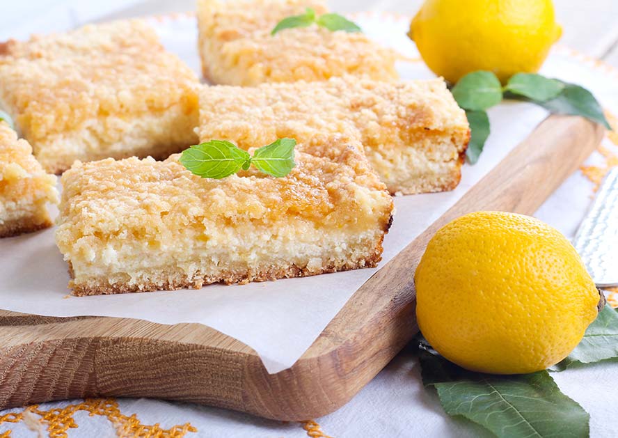 Lemon cheesecake bars topped with mint on a wooden tray with whole lemons