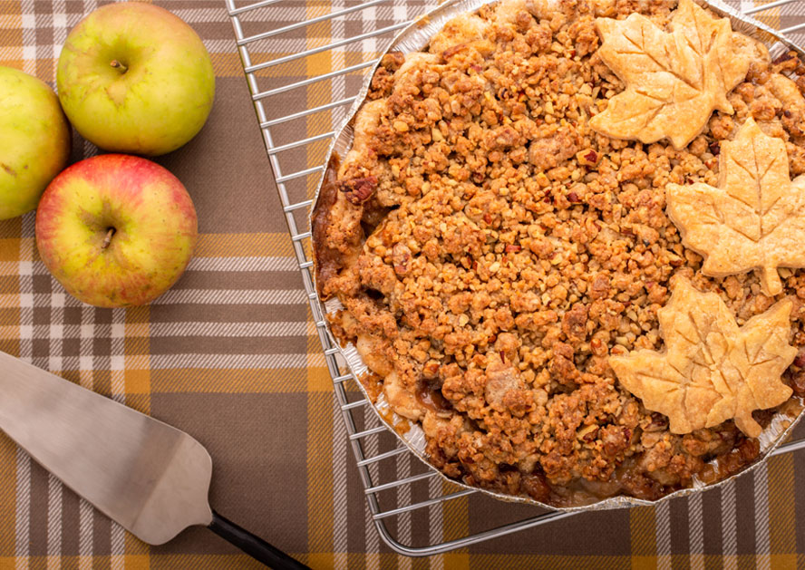Apple crumb pie with pastry leaves on a cooling rack, beside apples and a pie server