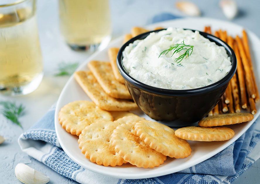 Bowl of dill dip on a plate with crackers, pretzel sticks, and glasses in the background