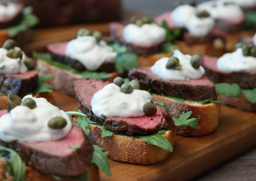 Sliced filet mignon appetizers on toasted bread with arugula, cream, and capers