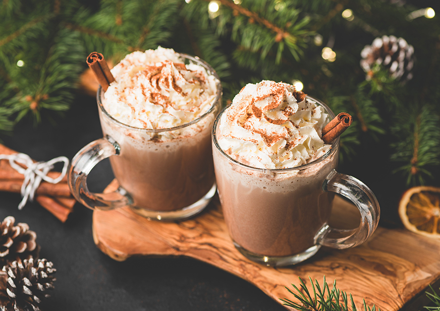 Two mugs of hot chocolate topped with whipped cream and cinnamon sticks on a wooden board