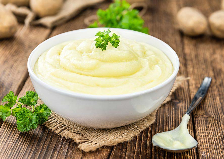 White bowl of mashed potatoes topped with parsley on a wooden table with a spoon
