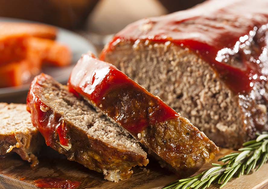 Sliced meatloaf with ketchup glaze on a wooden board, with carrots and rosemary nearby.