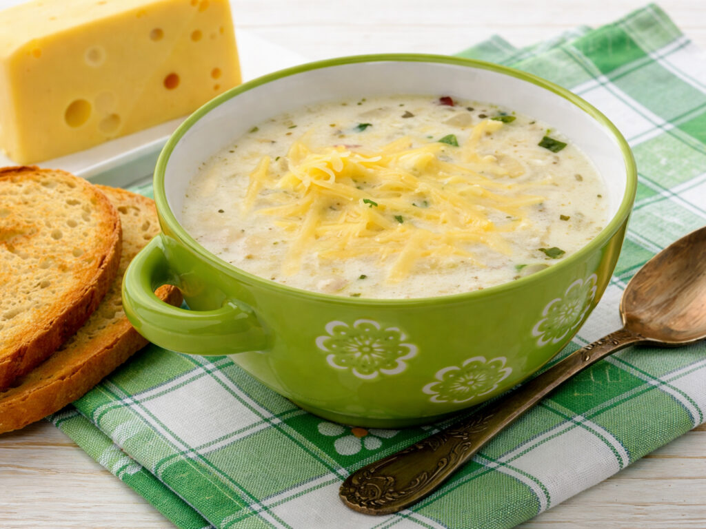 Bowl of creamy potato soup topped with shredded cheese, with bread slices and a spoon beside it.