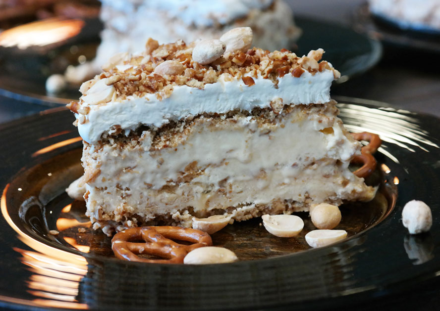 Slice of ice cream cake topped with whipped cream, peanuts, and pretzels on a black plate