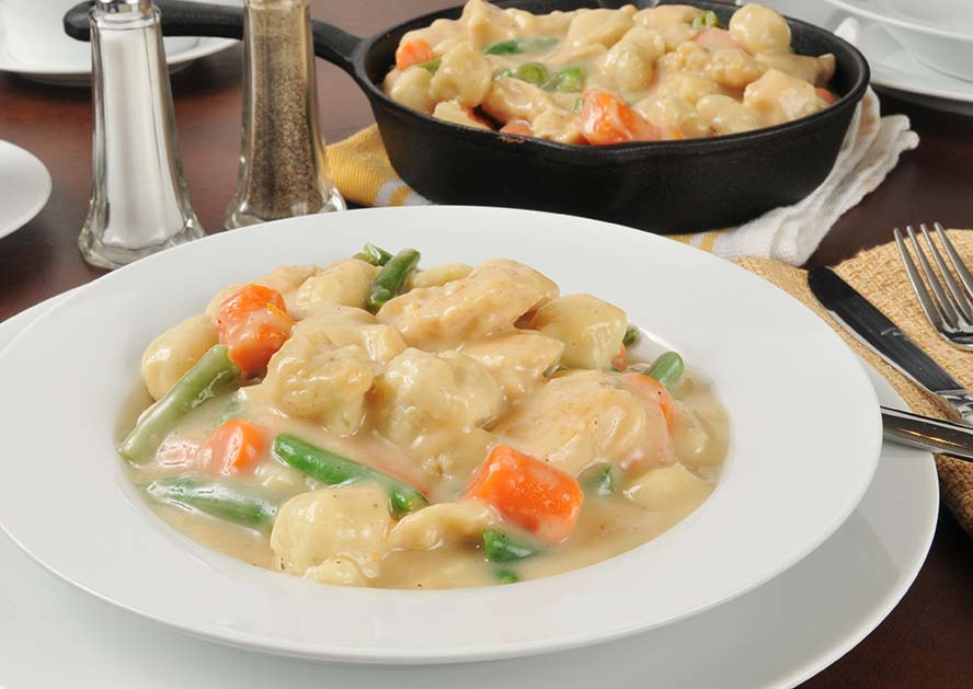Plate of chicken and dumplings with carrots and green beans, with a skillet in the background