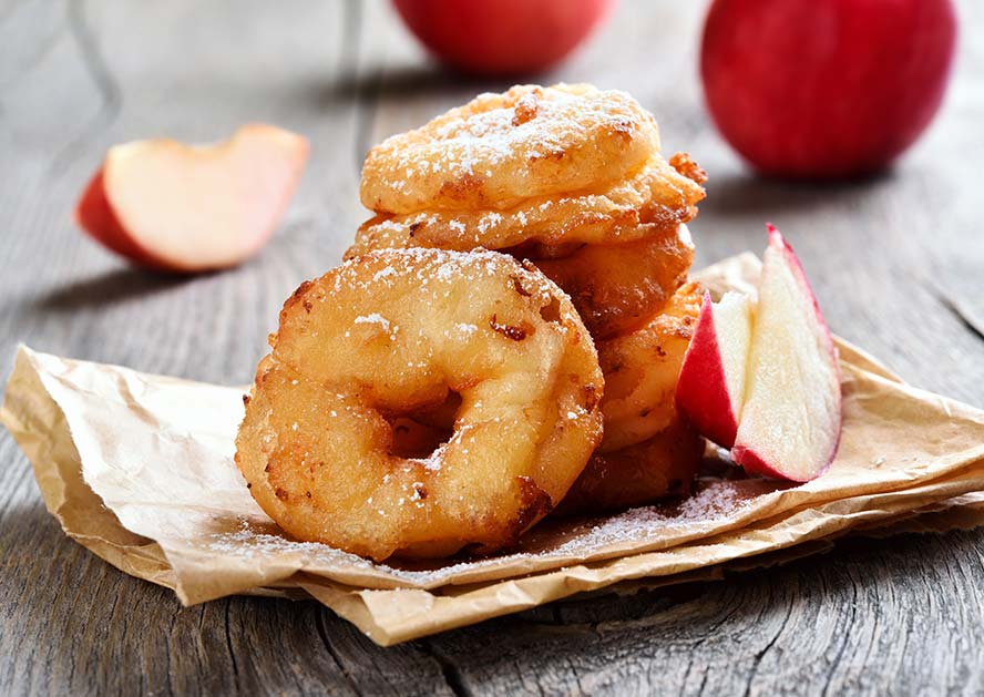 Stack of apple fritters dusted with powdered sugar on brown paper, with apple slices nearby