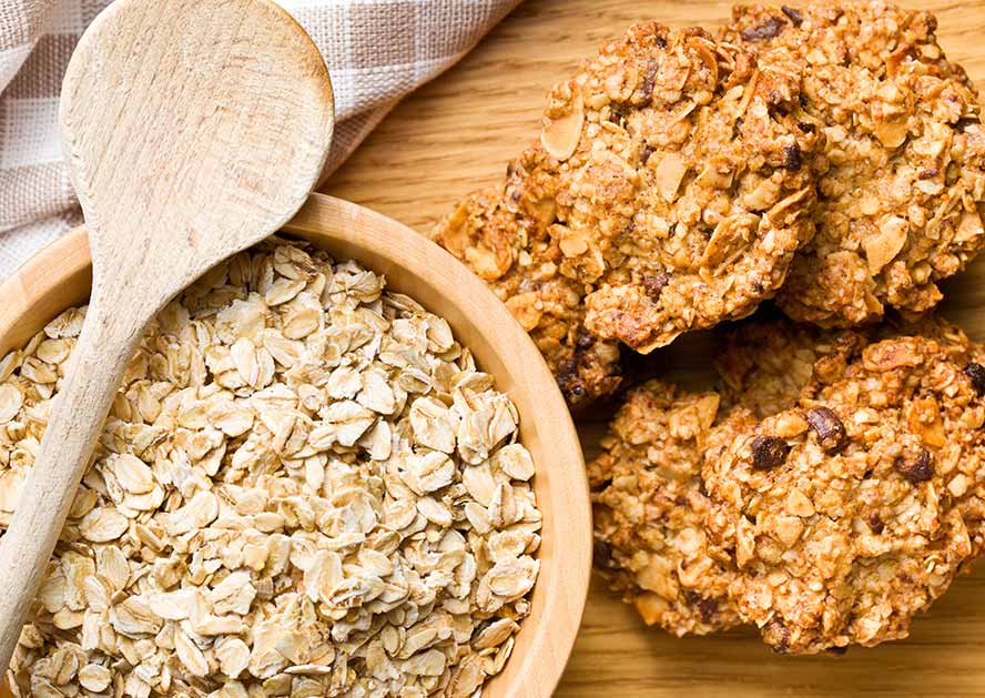 Bowl of rolled oats with a wooden spoon beside oatmeal cookies on a wooden surface