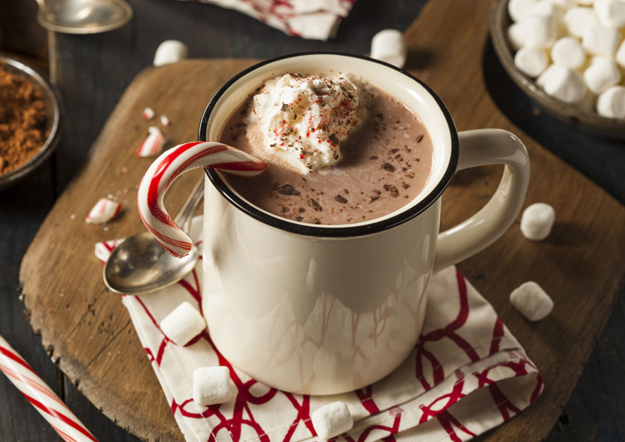 Mug of hot chocolate topped with whipped cream and a candy cane on a wooden board, with marshmallows nearby