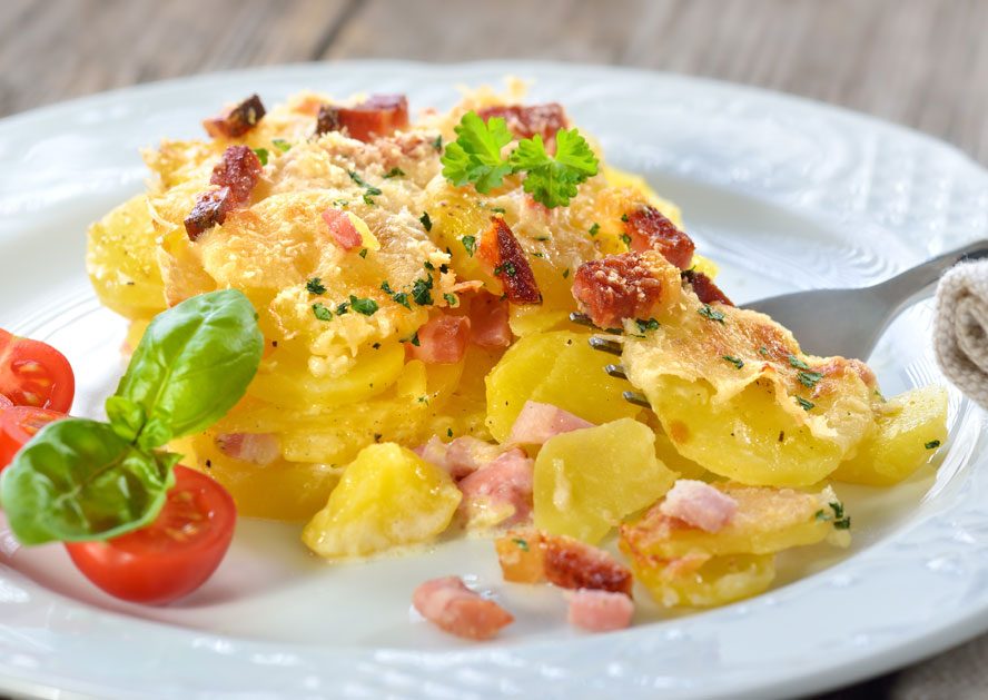 Baked cheesy potatoes with ham, herbs, cherry tomatoes, and basil on a white plate.