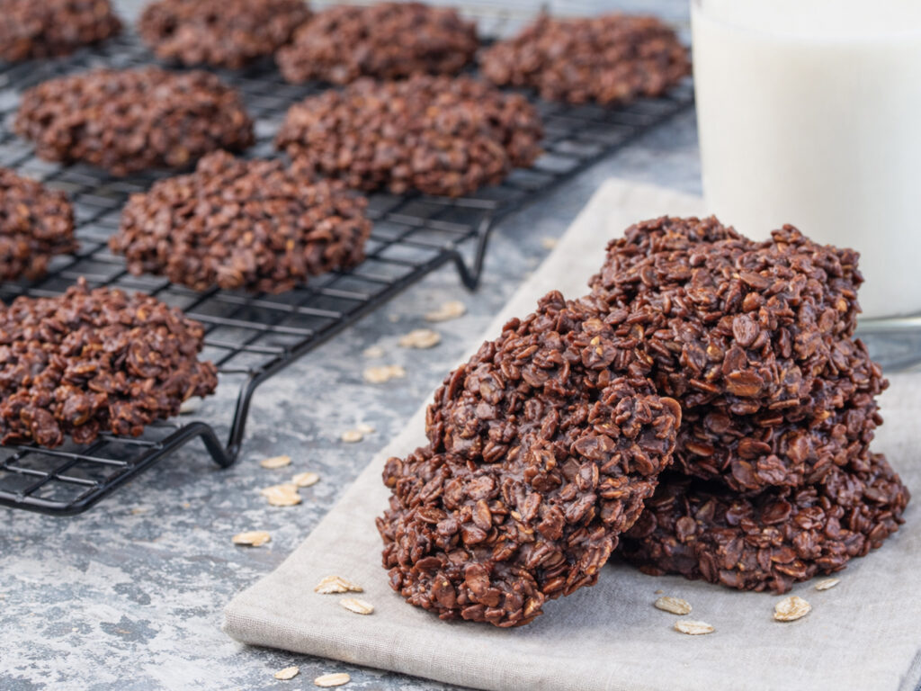 Stack of chocolate oatmeal no-bake cookies on a cloth, with more cooling on a rack and a glass of milk