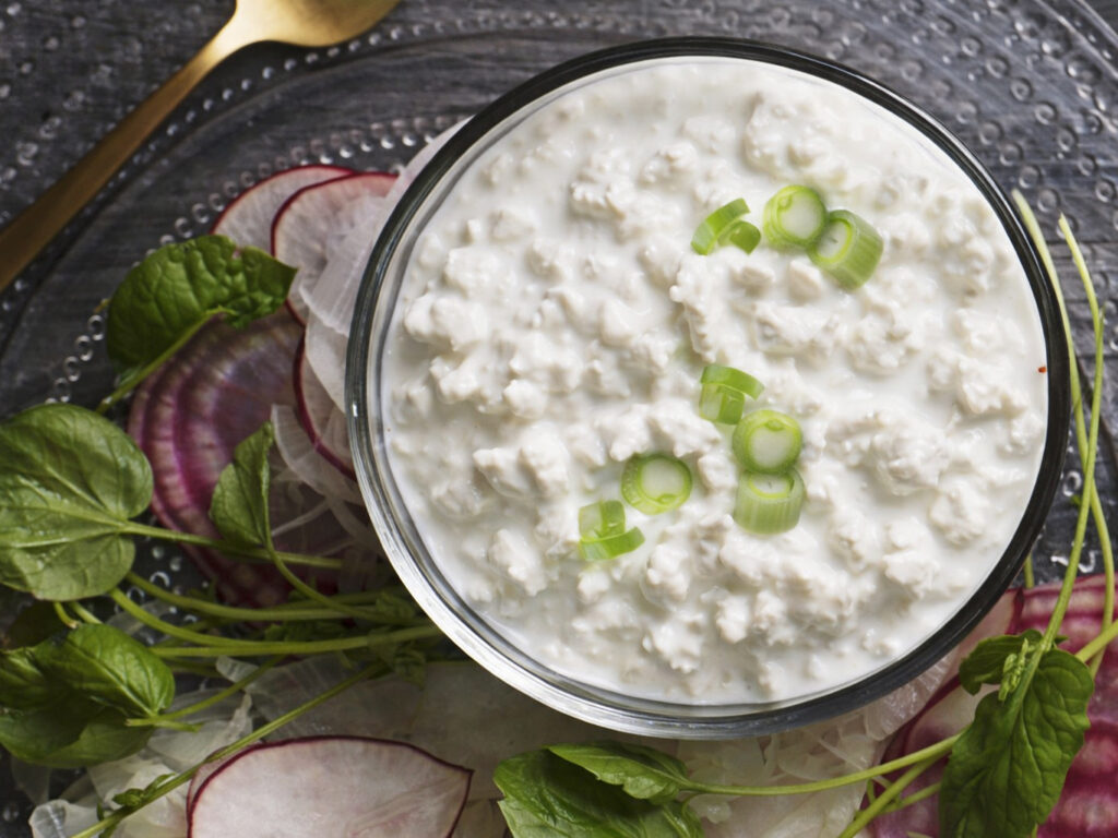 Bowl of cottage cheese topped with sliced green onions, surrounded by radish slices and leafy greens.