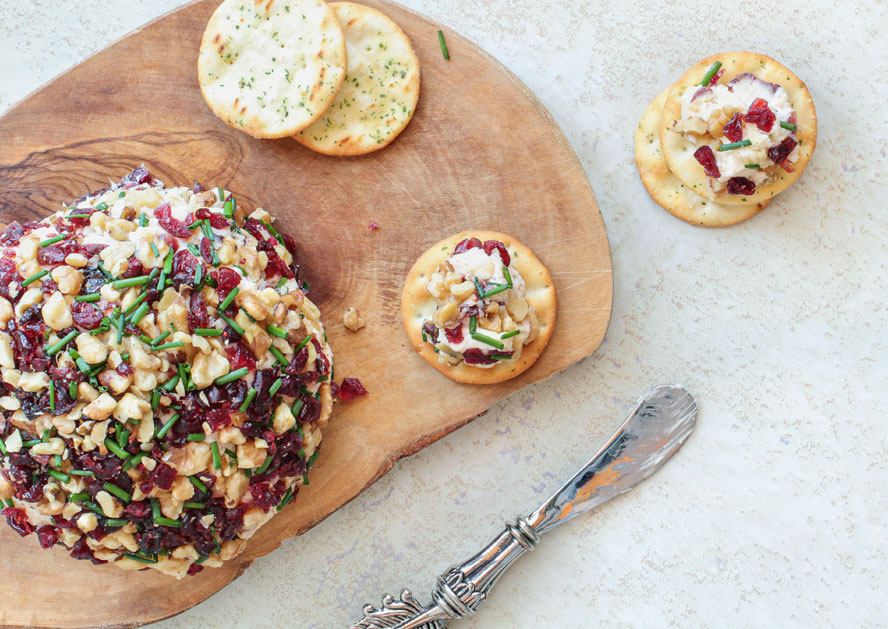 Cheese ball topped with bacon, cranberries, and chives on a wooden board with crackers