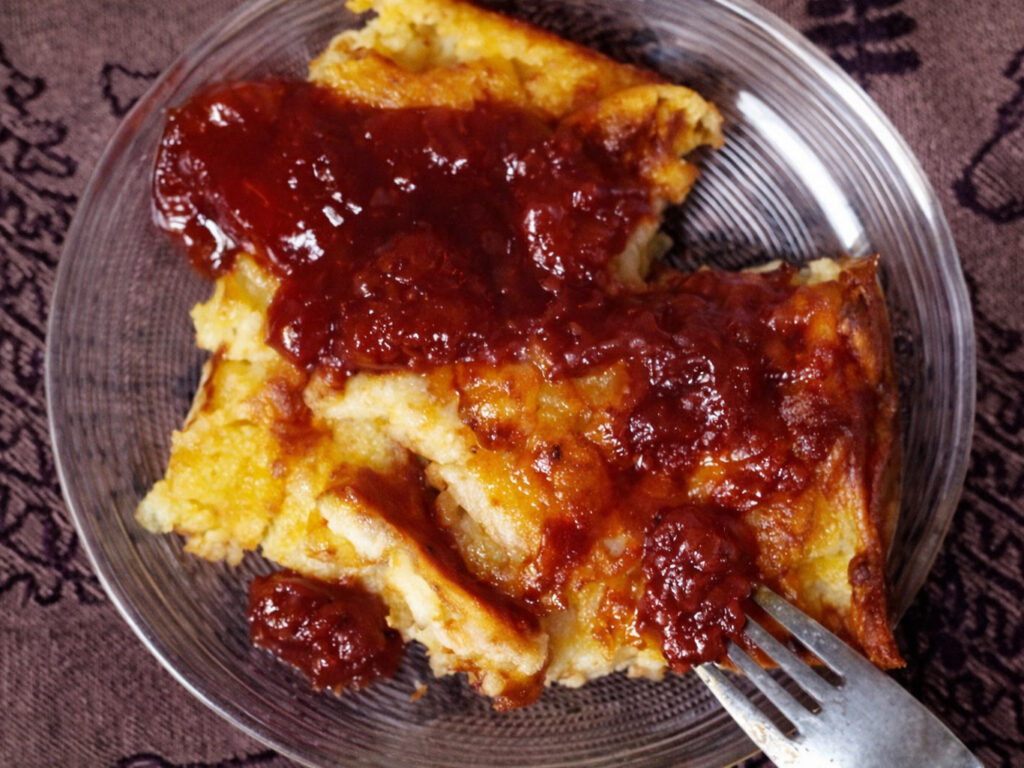 Slice of oven pancake topped with red berry sauce on a glass plate with a fork
