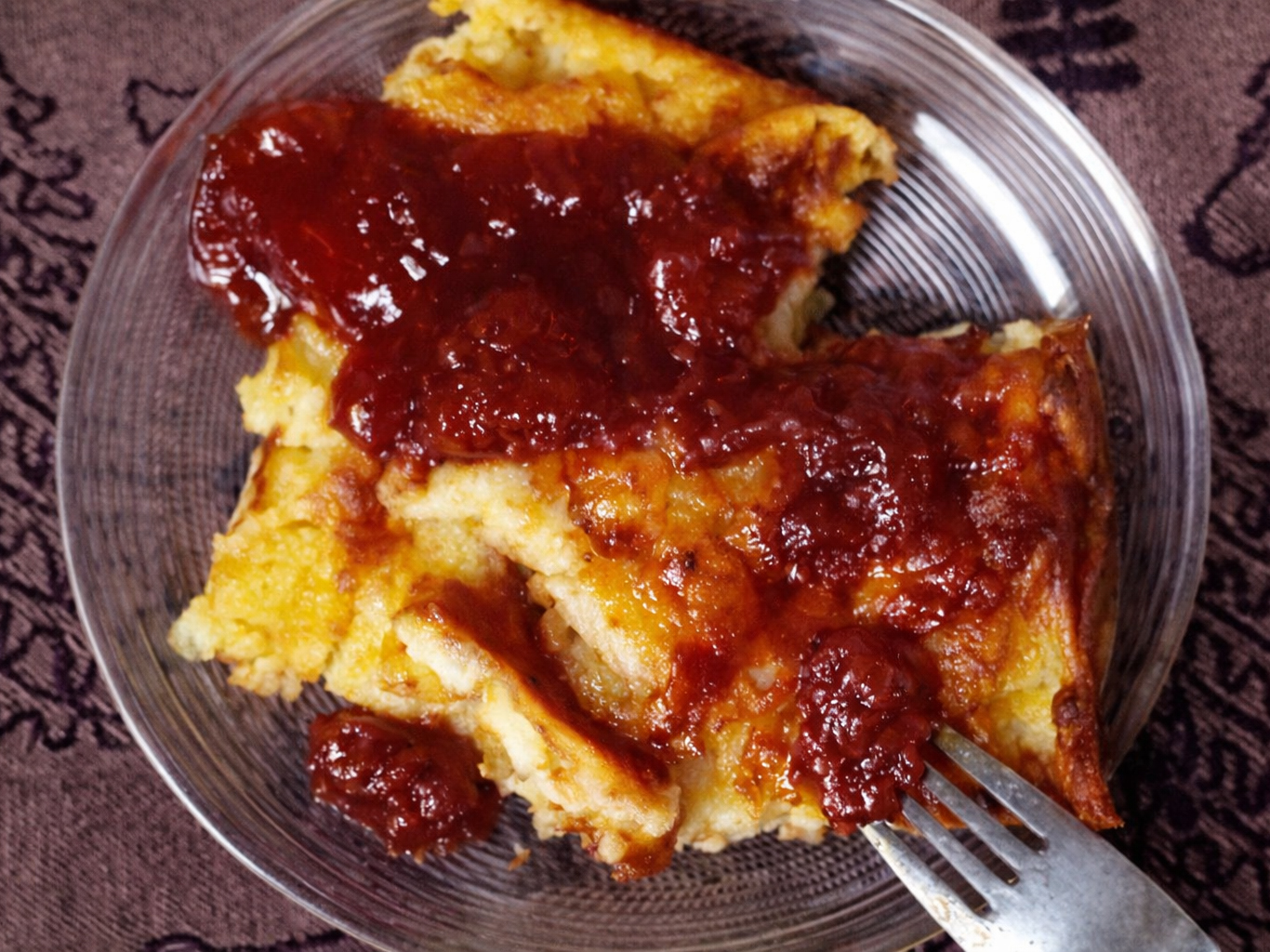 Slice of oven pancake topped with red berry sauce on a glass plate with a fork