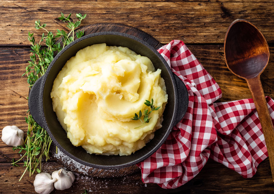 Bowl of mashed potatoes with thyme on a wooden table, with garlic, a spoon, and a red checkered cloth.