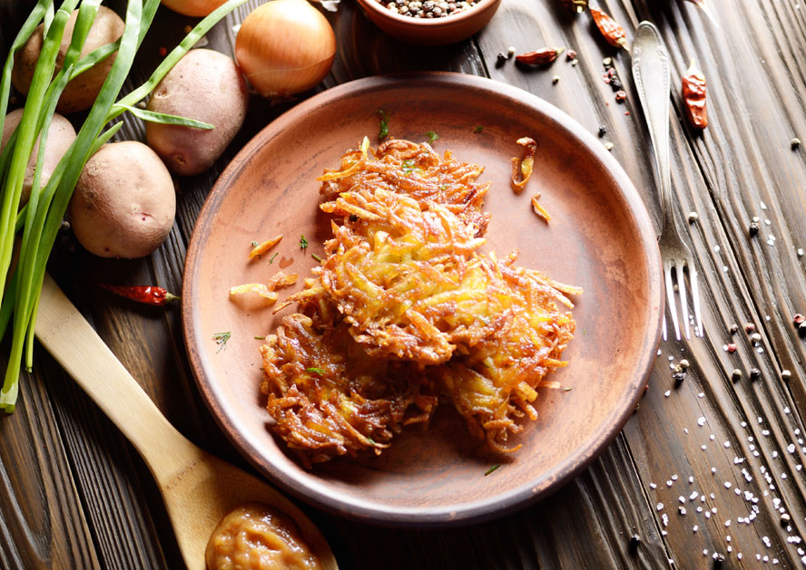 Plate of crispy potato latkes on a rustic table with potatoes, onions, and utensils