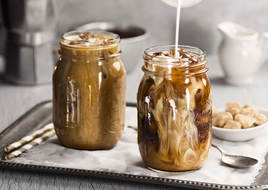 Two mason jars of iced coffee with cream being poured into one