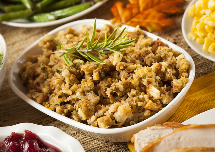 White bowl of bread stuffing garnished with rosemary, surrounded by side dishes