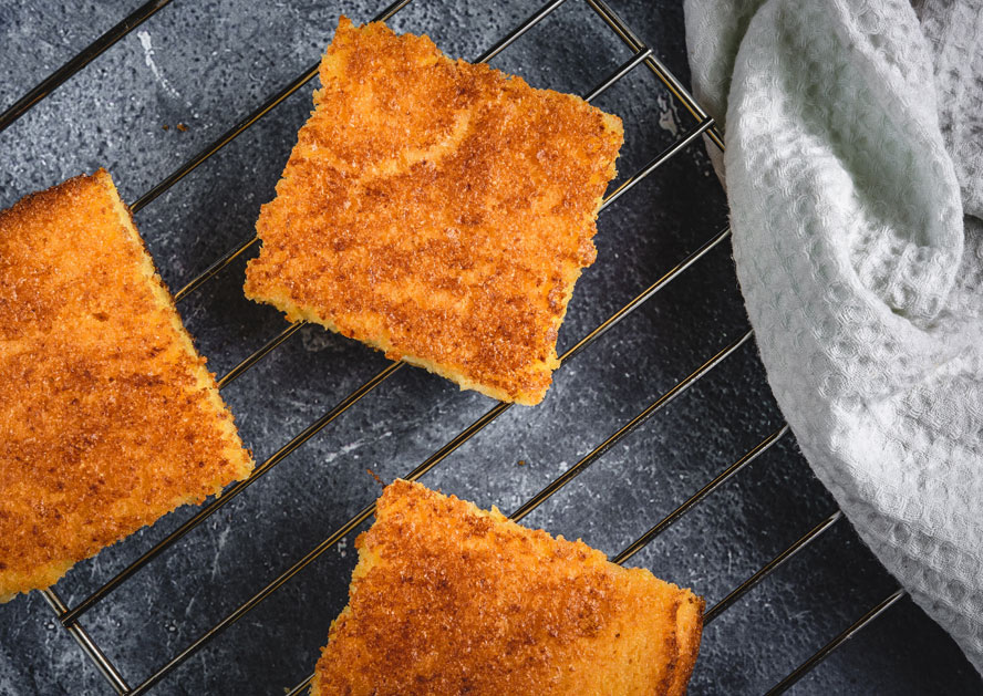 Square pieces of golden cornbread cooling on a wire rack beside a white cloth.