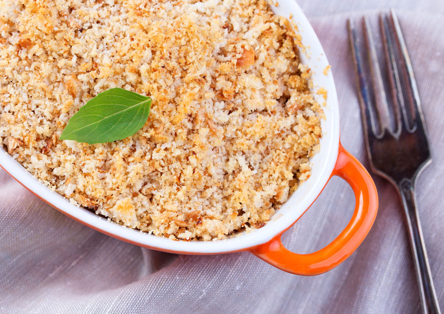 Baked casserole in a white dish with an orange handle, topped with breadcrumbs and a basil leaf, beside a fork.