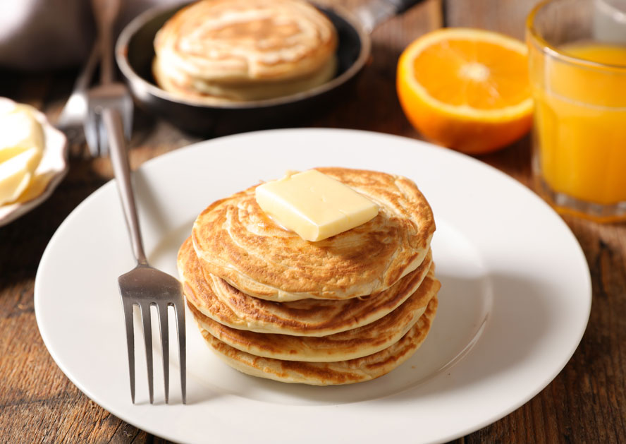 Stack of pancakes with butter on a plate, with a fork, orange slices, and a glass of orange juice