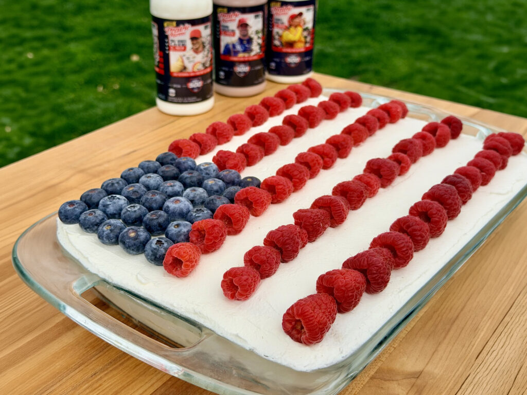 Rectangular ice cream cake topped with blueberries and raspberries arranged like an American flag, set in a glass dish with milk bottles in the background.