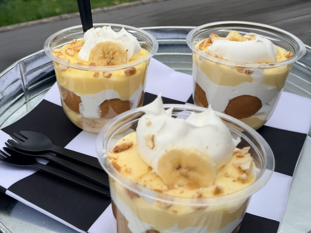 Three banana pudding cups layered with vanilla pudding, whipped cream, and cookies, topped with banana slices and cookie crumbles, served with plastic spoons on a tray.
