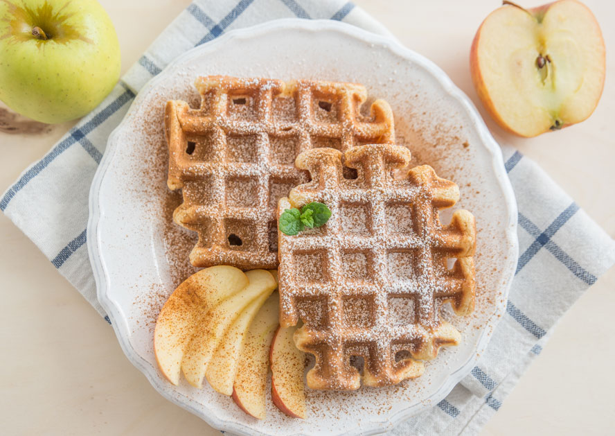 Two waffles on a plate, dusted with powdered sugar and cinnamon, with apple slices and whole apples nearby.