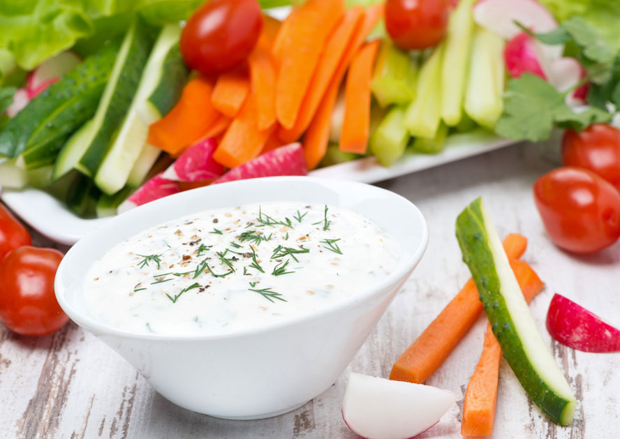 White bowl of creamy herb dip surrounded by sliced vegetables and cherry tomatoes