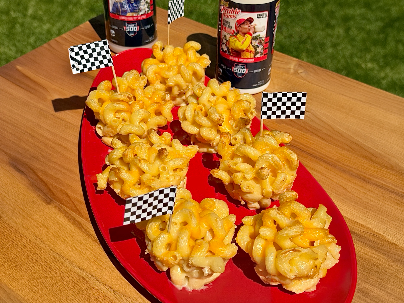 Baked mac and cheese cups arranged on a red platter with checkered racing flags, featuring golden crispy edges and creamy cheesy pasta, with Prairie Farms Milk bottles in the background.
