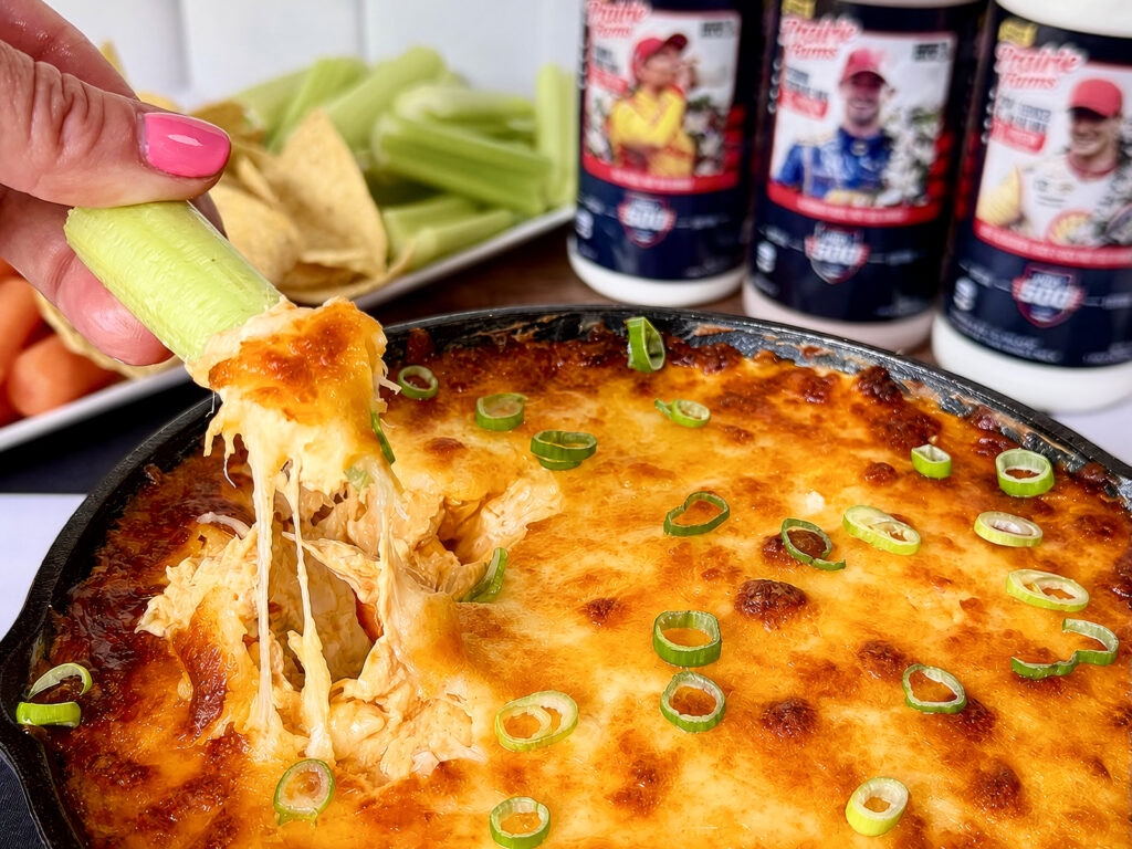 A cheesy baked buffalo chicken skillet dip topped with sliced green onions, as a celery stick lifts a gooey, melted portion, with chips, celery sticks and milk bottles in the background.